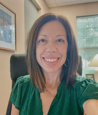Smiling woman in a green blouse seated in an office with windows and a lamp in the background