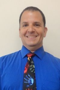Smiling person in a bright blue shirt and colorful tie against a plain wall