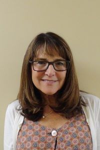 Smiling woman with glasses and shoulder-length brown hair against a plain beige wall