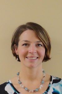 Portrait of a smiling person with short brown hair, wearing a beaded necklace against a beige background