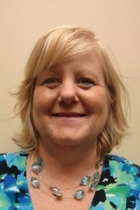 Smiling woman with blonde hair wearing a blue patterned top and a silver necklace against a beige wall