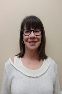 Smiling person with dark hair and glasses wearing a white sweater against a beige wall