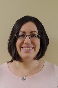 Smiling person with glasses and a bob haircut, wearing a light pink top against a plain beige background