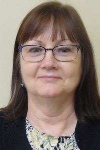 Portrait of a woman wearing glasses and a black cardigan, facing forward against a beige background