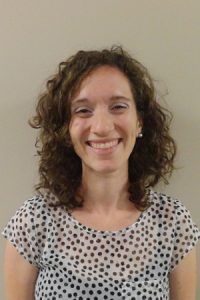 Smiling person with curly hair wearing a white polka-dot shirt against a plain background