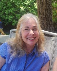 Smiling person in a blue shirt seated outdoors beside a tree and greenery.