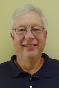 Smiling man with glasses and gray hair wearing a dark polo shirt against a plain wall