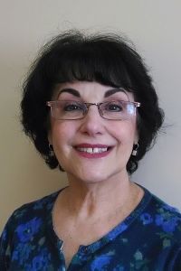 Smiling person with dark hair and glasses wearing a blue patterned top against a plain background