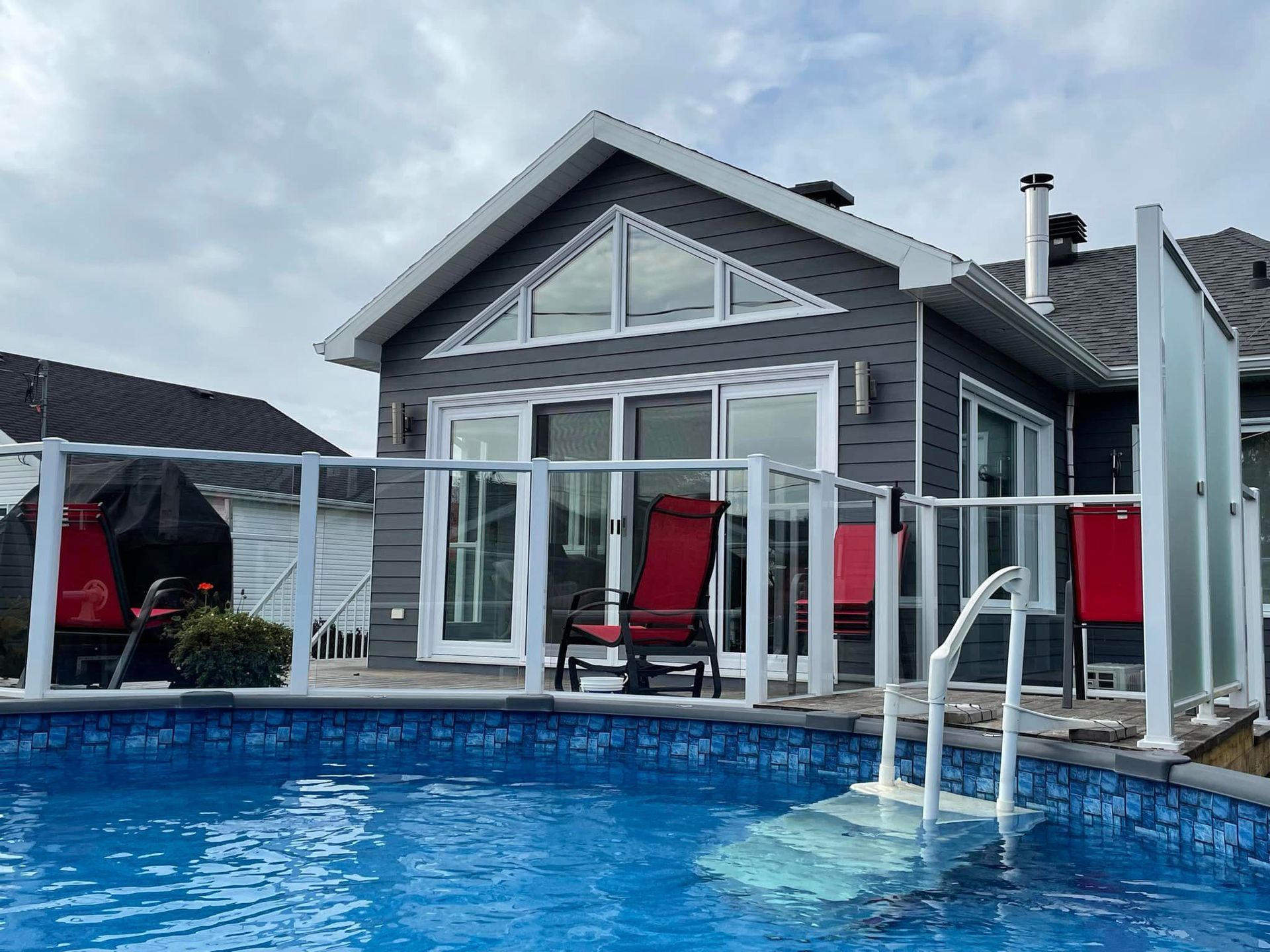Au premier plan, une piscine creusée ; à l'arrière-plan, une maison grise avec des balustrades blanches et deux chaises rouges.