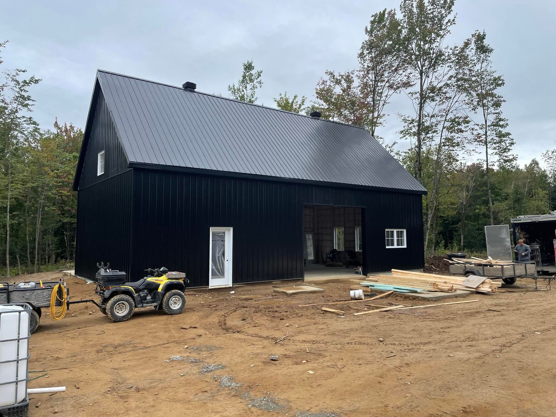 Un hangar moderne noir à poteaux, avec un toit en métal et une porte blanche, situé dans une zone boisée, avec un quad garé à proximité.