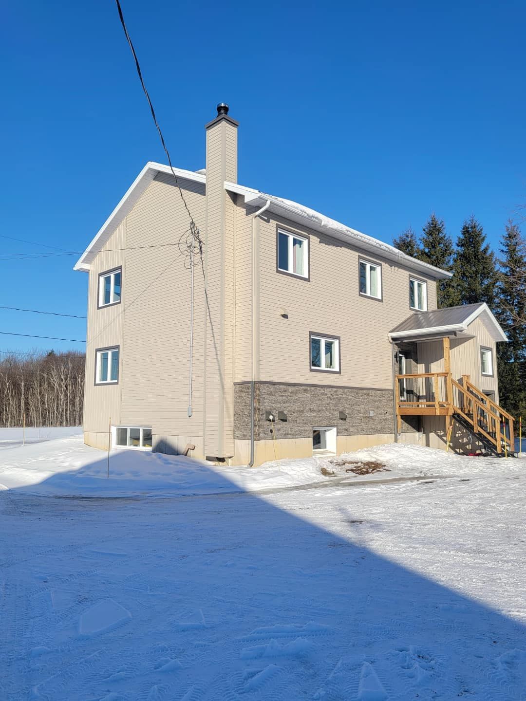 Maison beige à deux étages avec des fondations en pierre, une cheminée et un porche en bois, entourée de neige sous un ciel bleu clair.