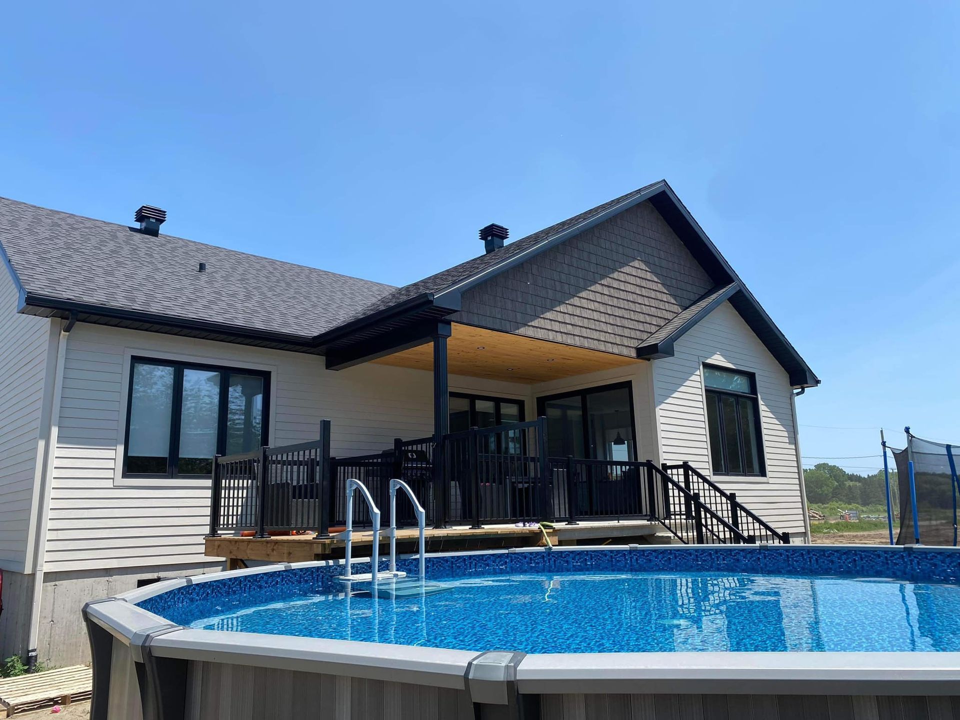 Une maison aux tons clairs, au toit sombre et à la terrasse couverte, vue depuis l'arrière d'une piscine hors sol sous un ciel bleu.
