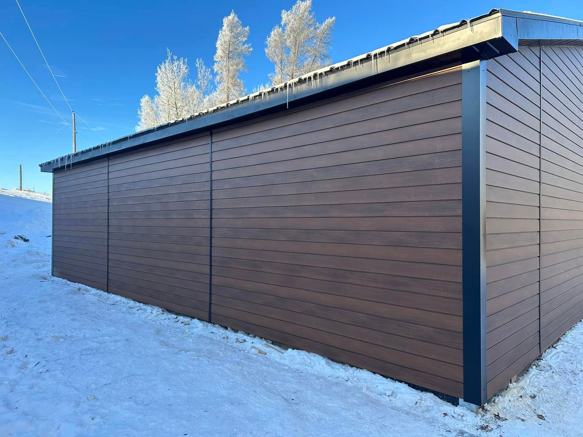 Un bâtiment de stockage aux panneaux horizontaux bruns se dresse dans un paysage enneigé, sous un ciel d'un bleu limpide.