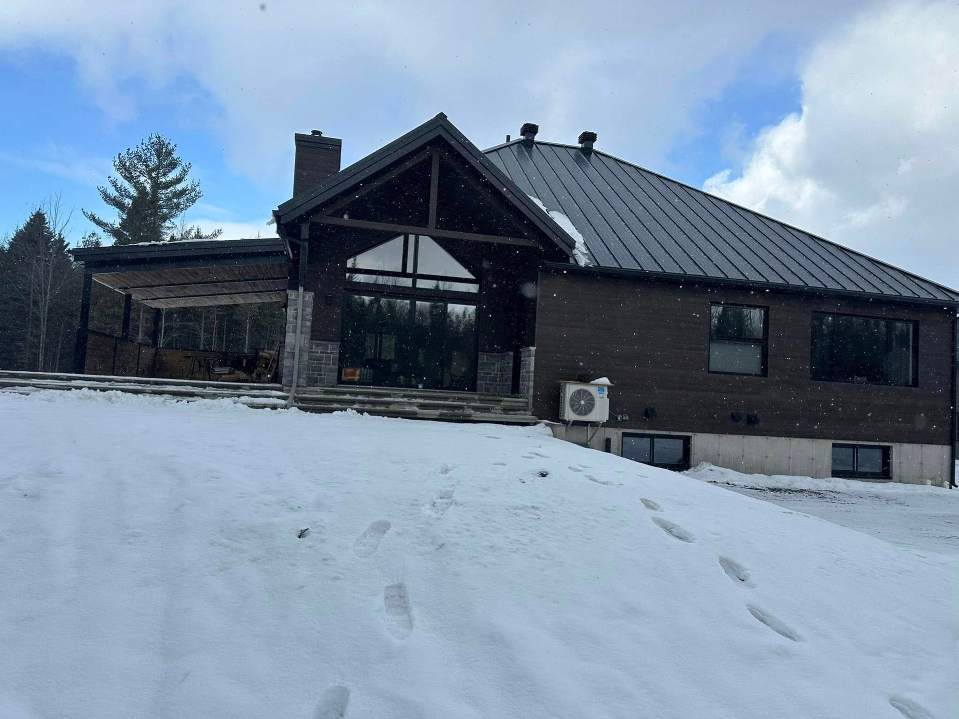 Une maison en briques sombres, au toit à pignon et avec un patio ouvert, se dresse dans un paysage enneigé sous un ciel bleu parsemé de légers nuages.