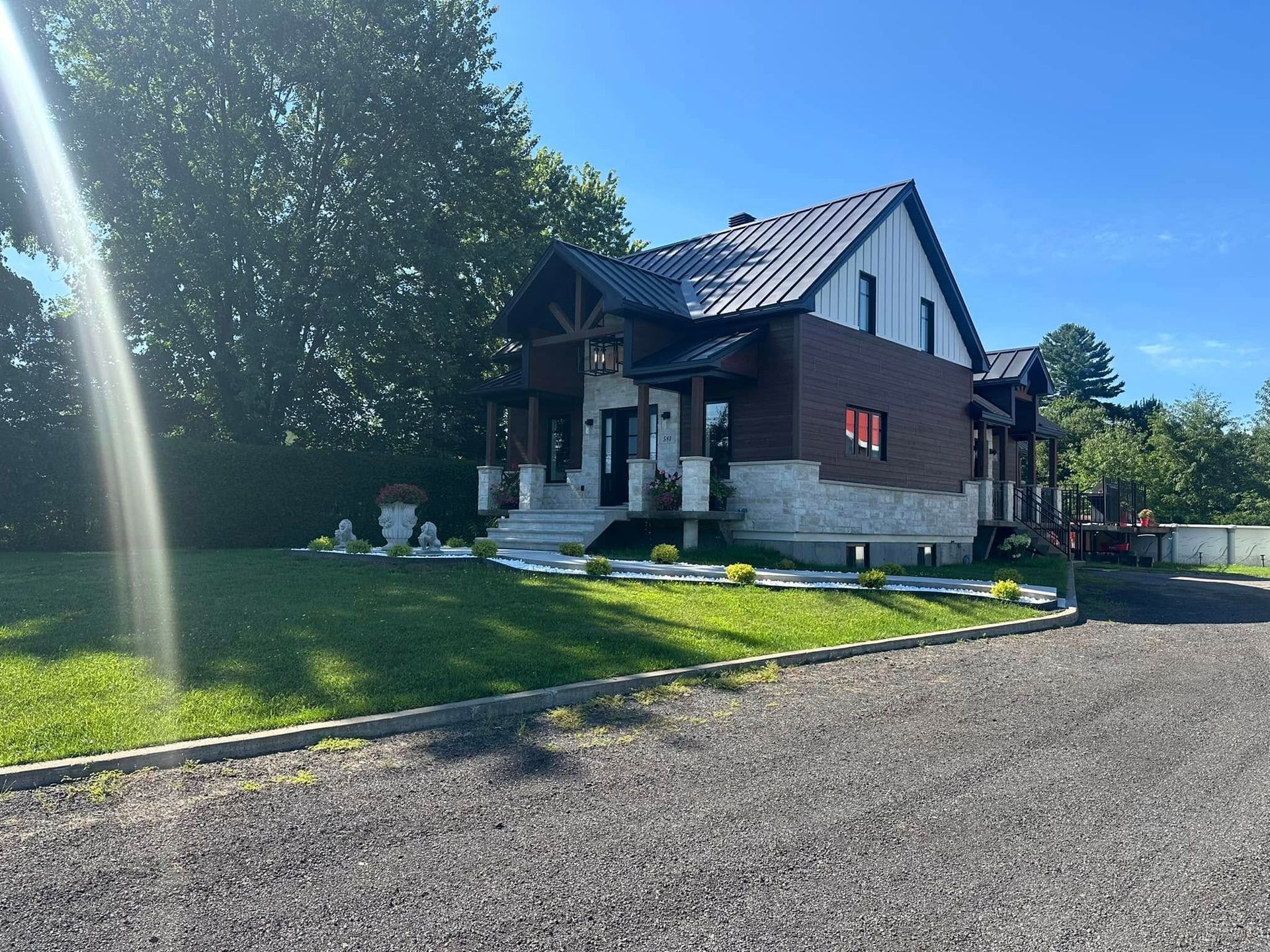 Une maison moderne avec un soubassement en pierre, un étage supérieur en briques sombres et un toit en métal, vue depuis une allée de gravier par une journée ensoleillée.