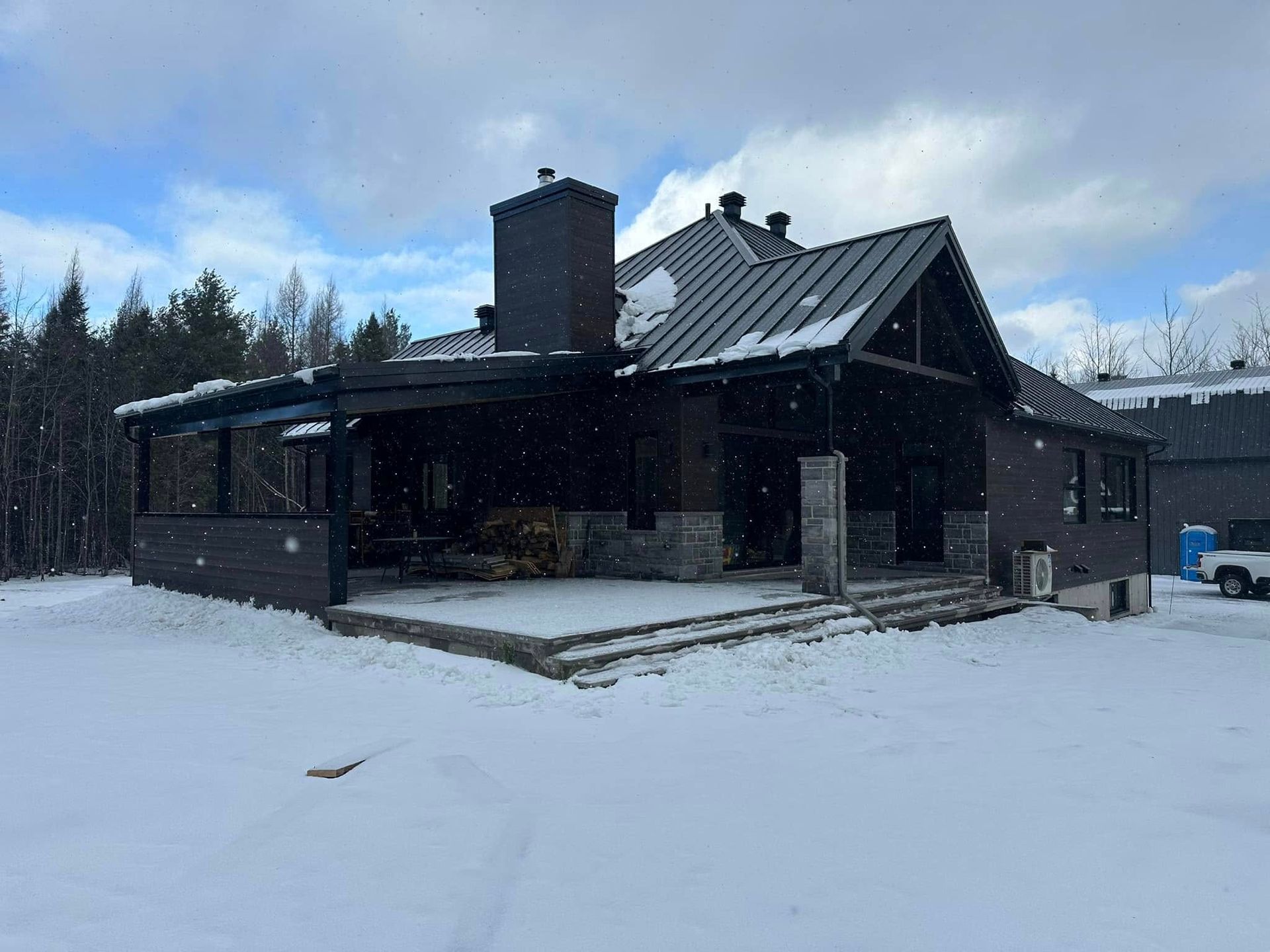 Une maison moderne brun foncé avec une cheminée en pierre et une grande terrasse couverte, entourée de neige sous un ciel nuageux.