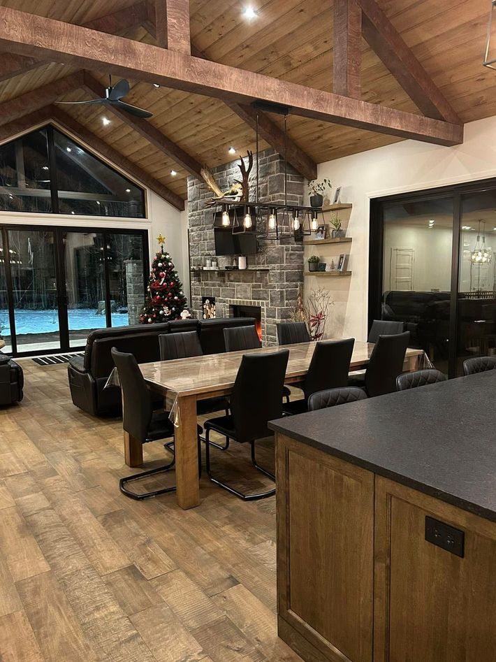Une salle à manger spacieuse et moderne à aire ouverte avec une cheminée en pierre, des poutres en bois, une grande table et des chaises en cuir foncé.