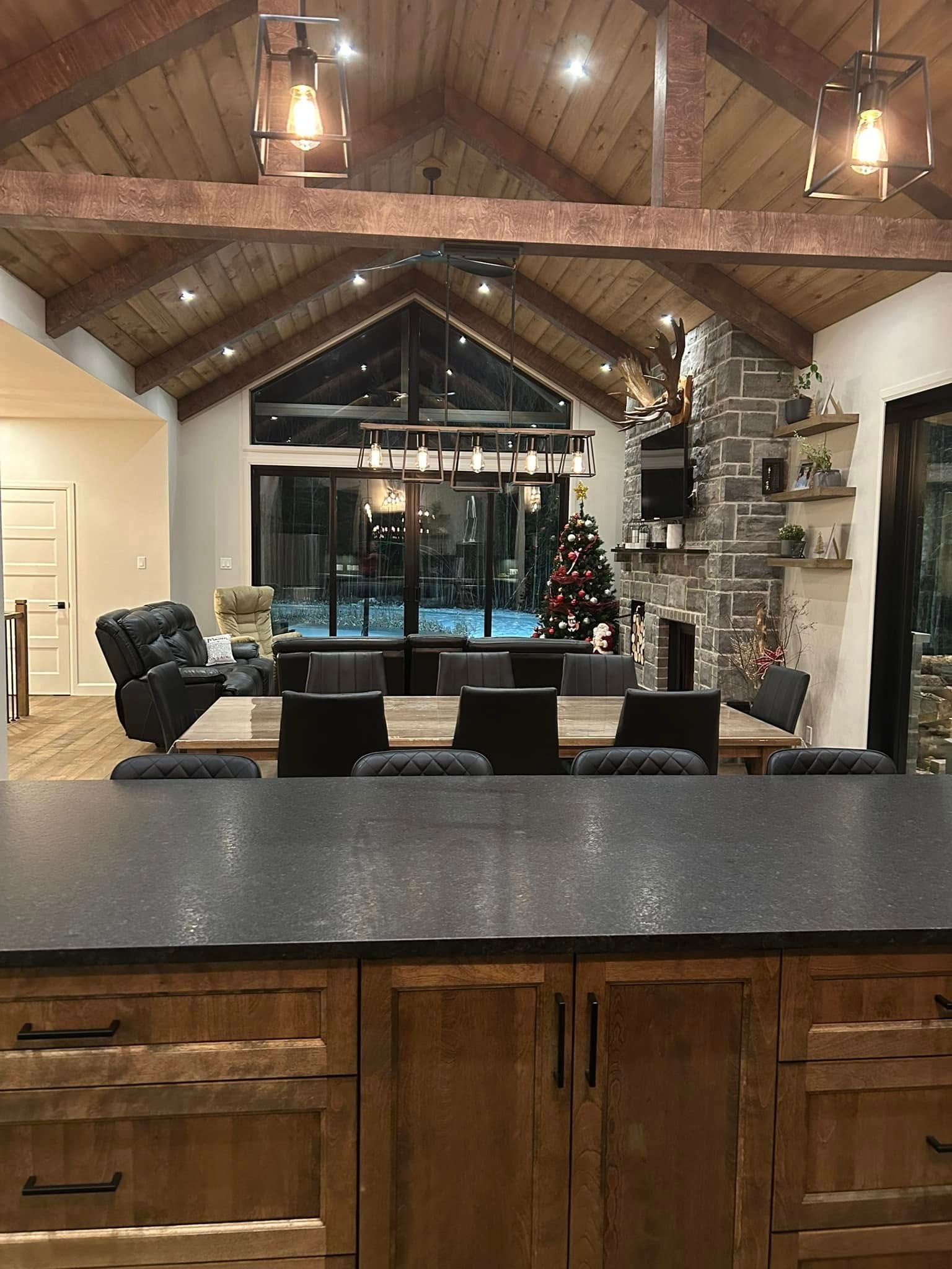 Cette pièce rustique à aire ouverte présente des plafonds voûtés en bois, une cheminée en pierre, une table à manger et un îlot de cuisine sombre.