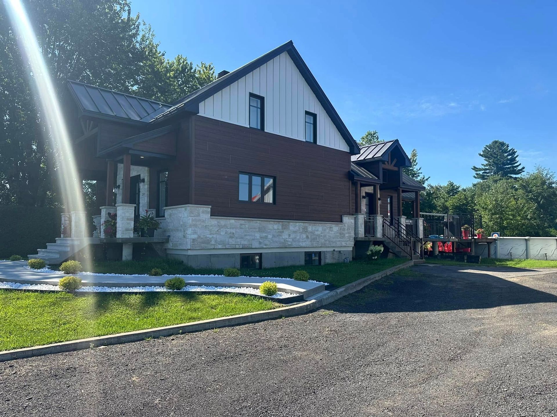 Une maison moderne avec des façades en briques et un bardage blanc, des fondations en pierre, un toit en métal et une allée en gravier, par une journée ensoleillée.