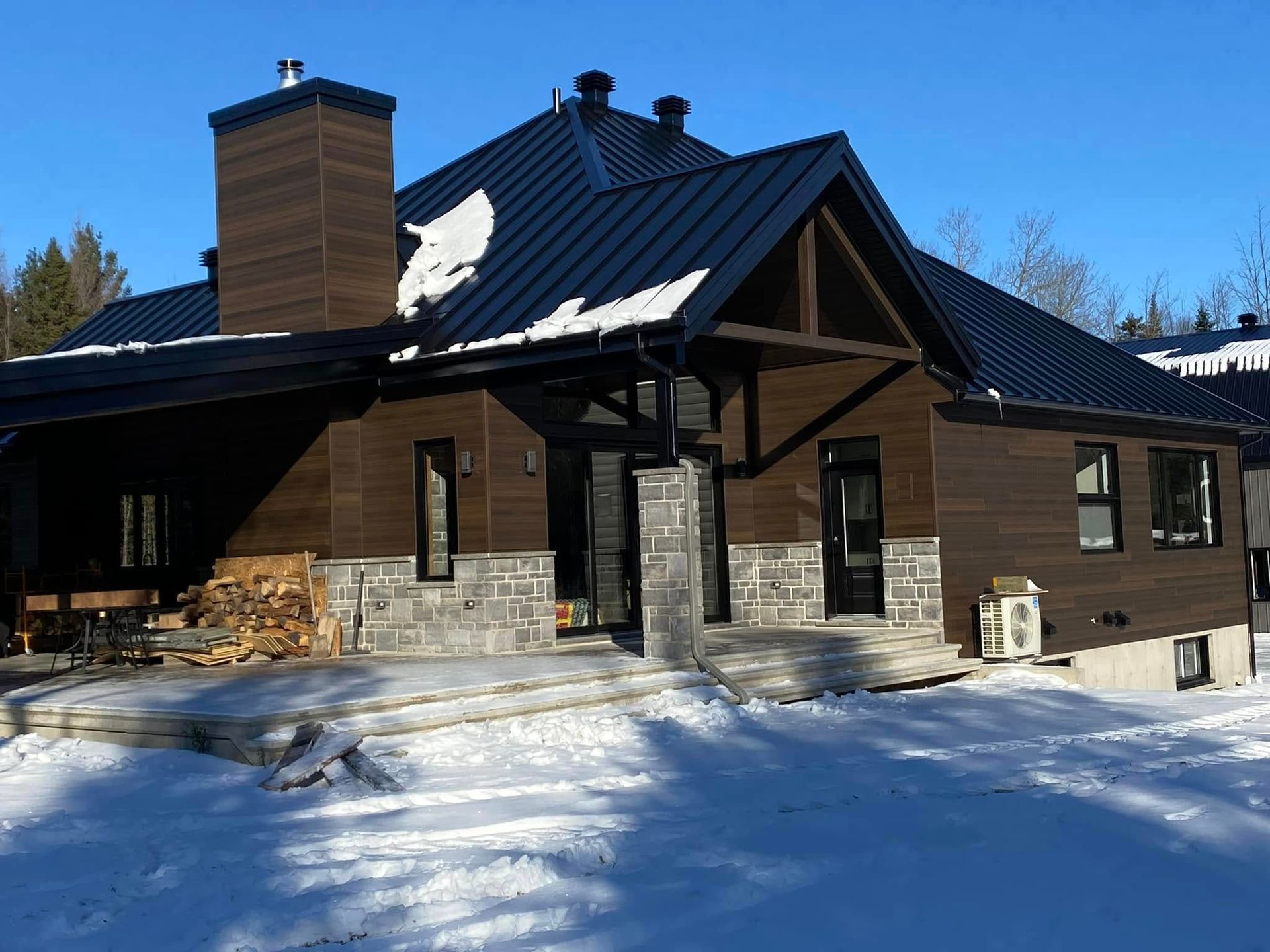 Une maison en bois sombre, avec des fondations en pierre et un toit en métal noir, entourée de neige sous un ciel bleu azur.