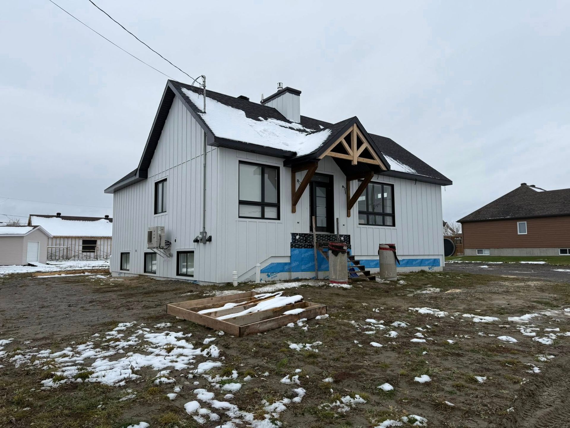 Une maison blanche en construction sur un terrain enneigé, avec un bardage vertical et un toit sombre, dont l'entrée principale est inachevée.