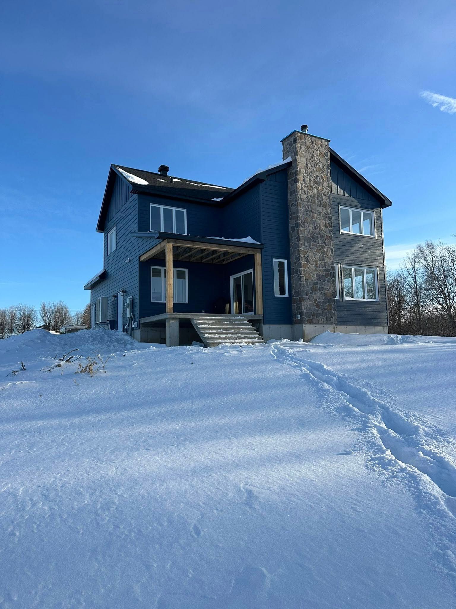 Une maison bleue à deux étages avec une cheminée en pierre se dresse dans un paysage enneigé sous un ciel d'un bleu limpide.