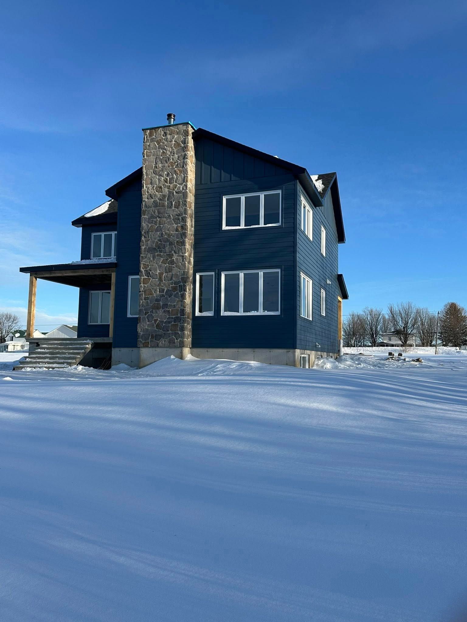 Une maison bleue à deux étages avec une cheminée en pierre se dresse dans une cour enneigée sous un ciel d'un bleu limpide.