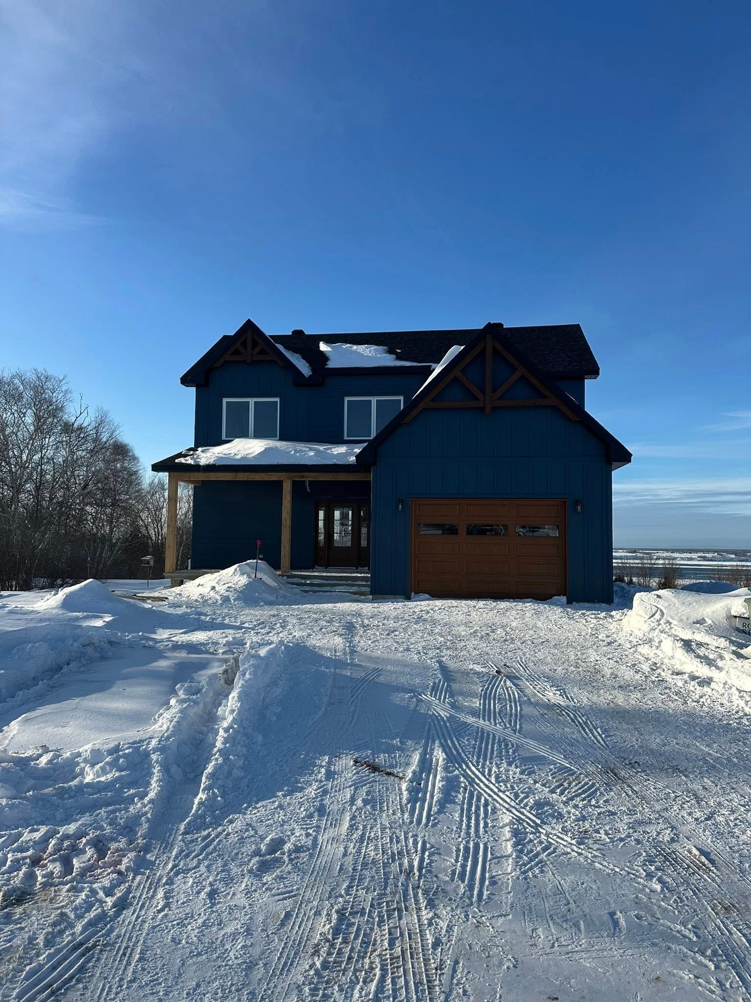 Une maison bleue à deux étages, avec une porte de garage en bois, se dresse dans un paysage enneigé sous un ciel d'un bleu limpide.
