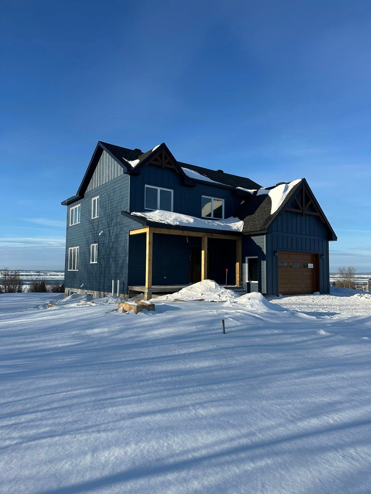 Une maison moderne de deux étages, d'un bleu foncé, en construction, entourée d'une épaisse couche de neige sous un ciel d'un bleu limpide.