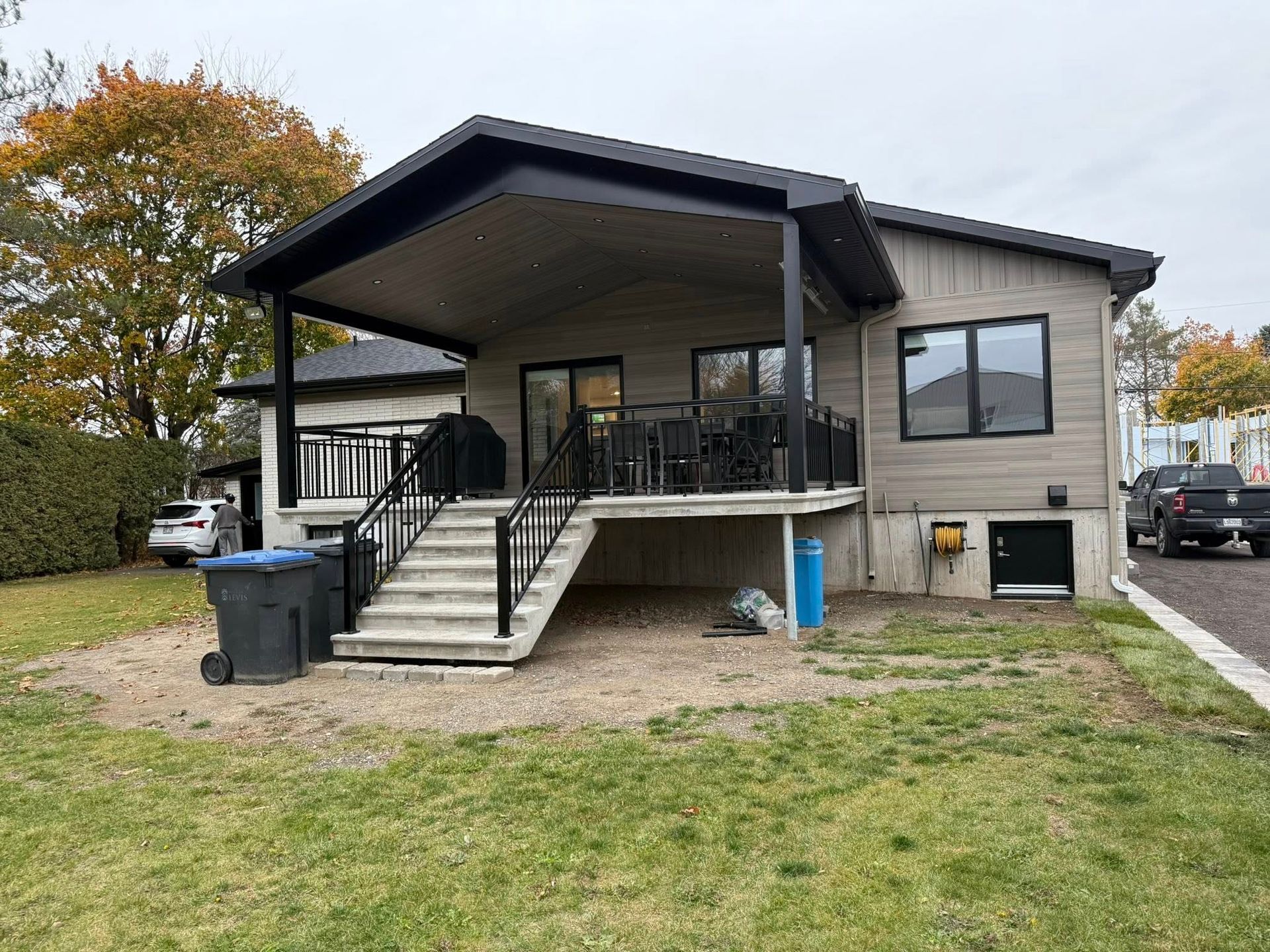 L'extérieur d'une maison moderne avec une terrasse couverte, un escalier en pierre menant à une cour arrière et un toit sombre sous un ciel couvert.