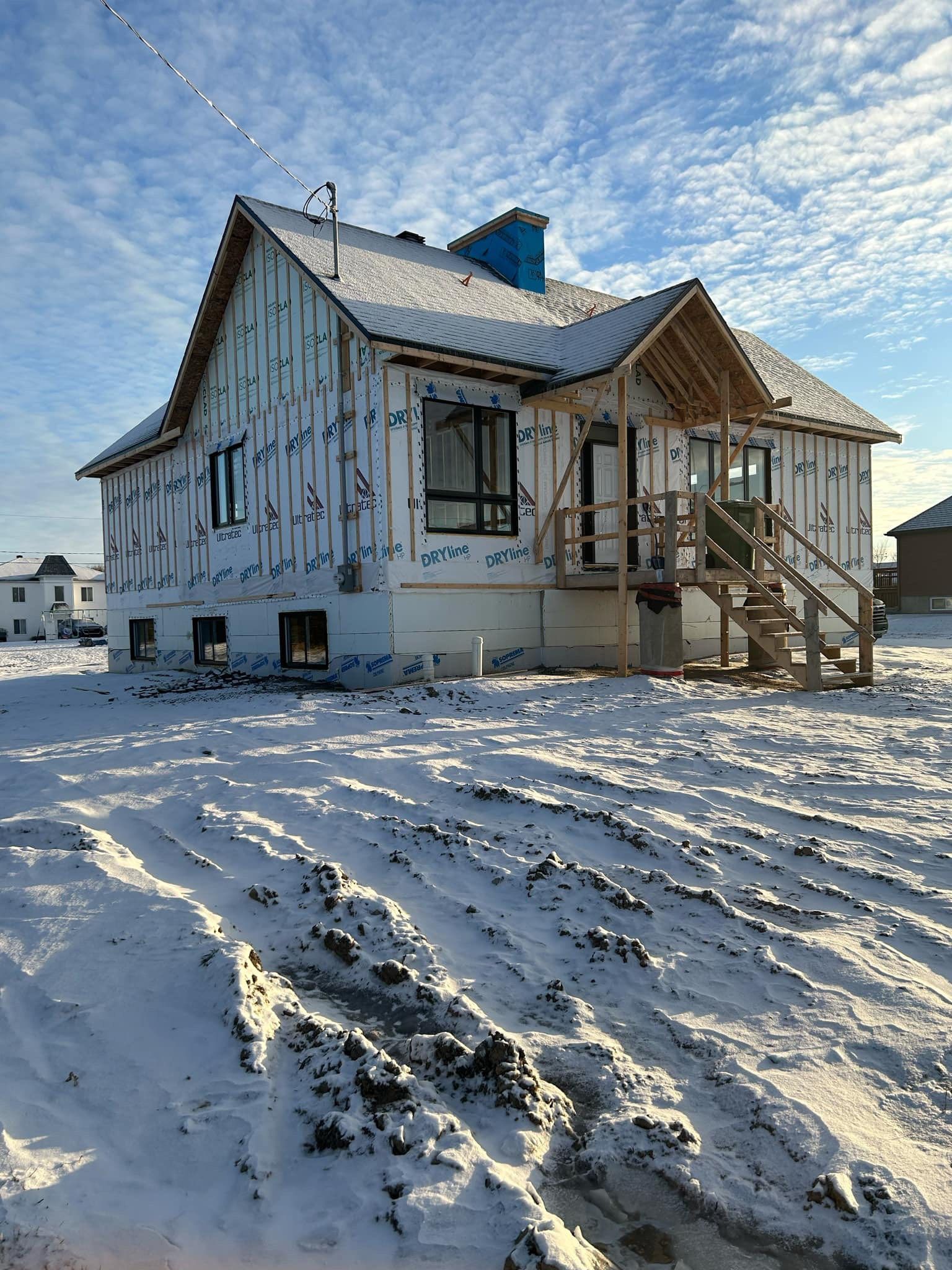 Une maison en construction, avec des panneaux extérieurs blancs et un encadrement d'entrée en bois, entourée de neige par une journée ensoleillée.