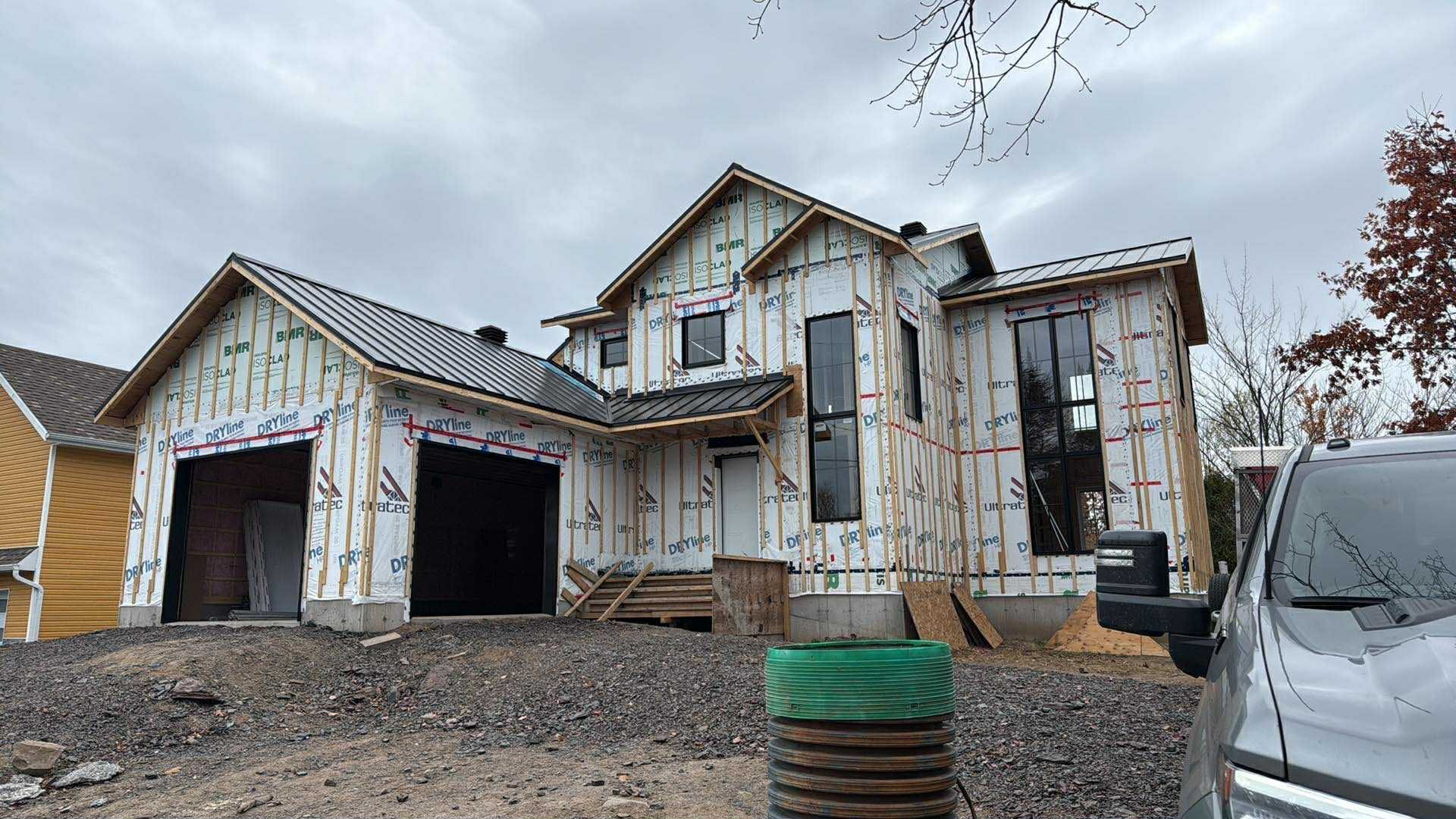 Une maison à deux étages en construction, avec un revêtement extérieur blanc, des cadres de fenêtres noirs et un garage inachevé.