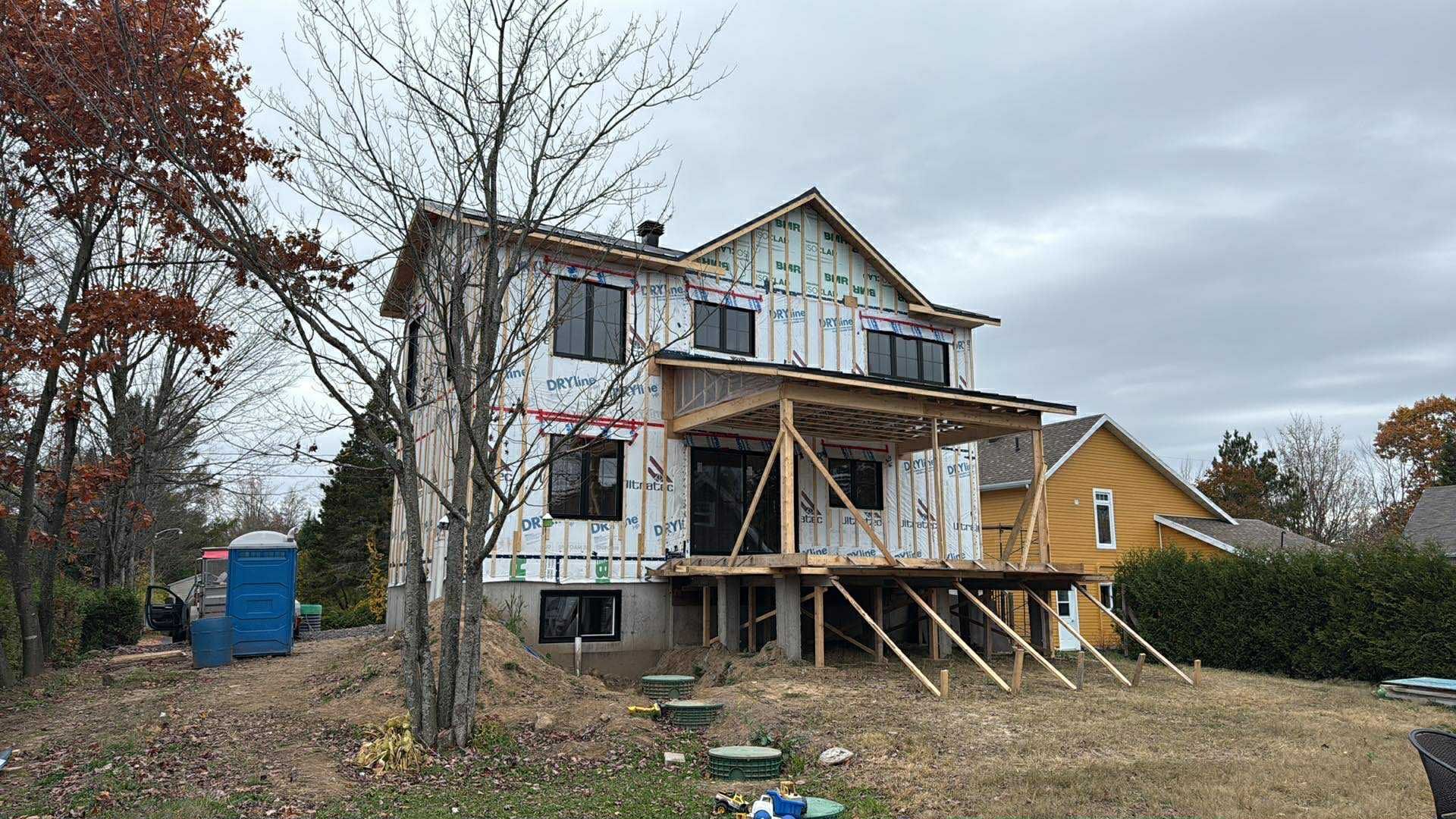 Une maison à plusieurs étages en construction, avec une charpente en bois apparente et une terrasse, située dans une cour arborée avec un abri mobile.