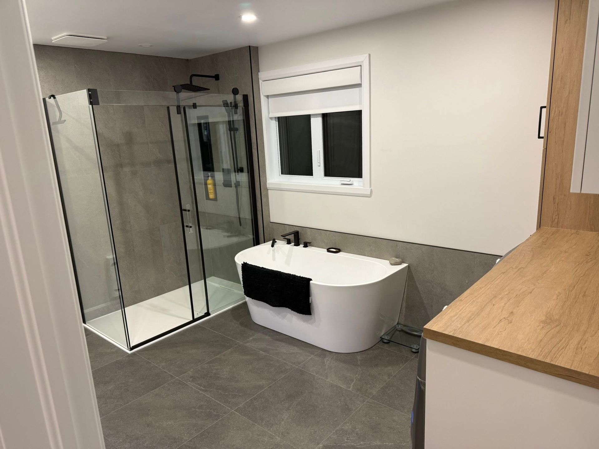 Salle de bains moderne avec douche vitrée, baignoire îlot blanche, carrelage gris au sol et meuble-lavabo en bois.