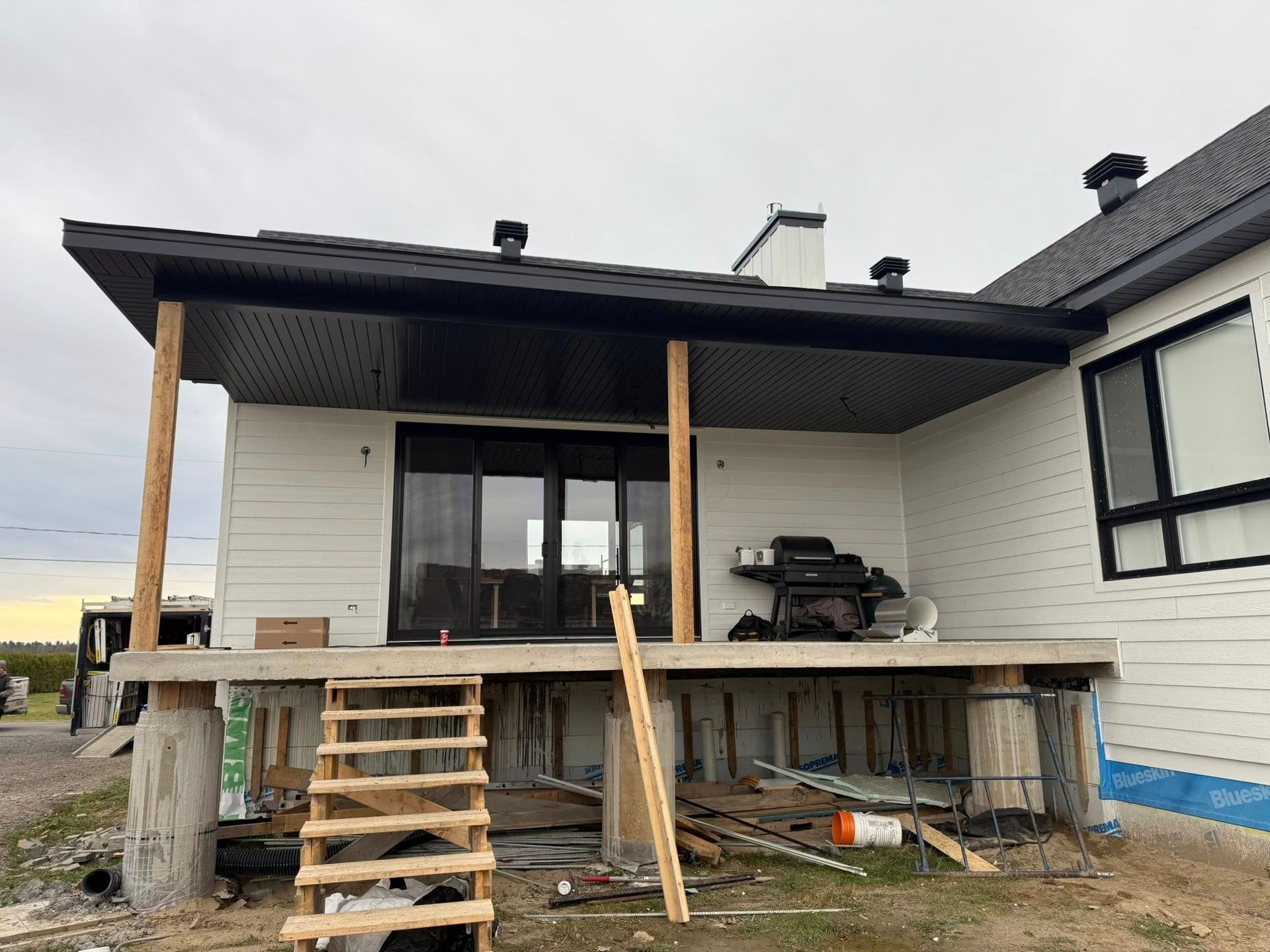 Une maison blanche avec une terrasse couverte, des piliers de soutien en bois et un escalier en béton inachevé en construction.