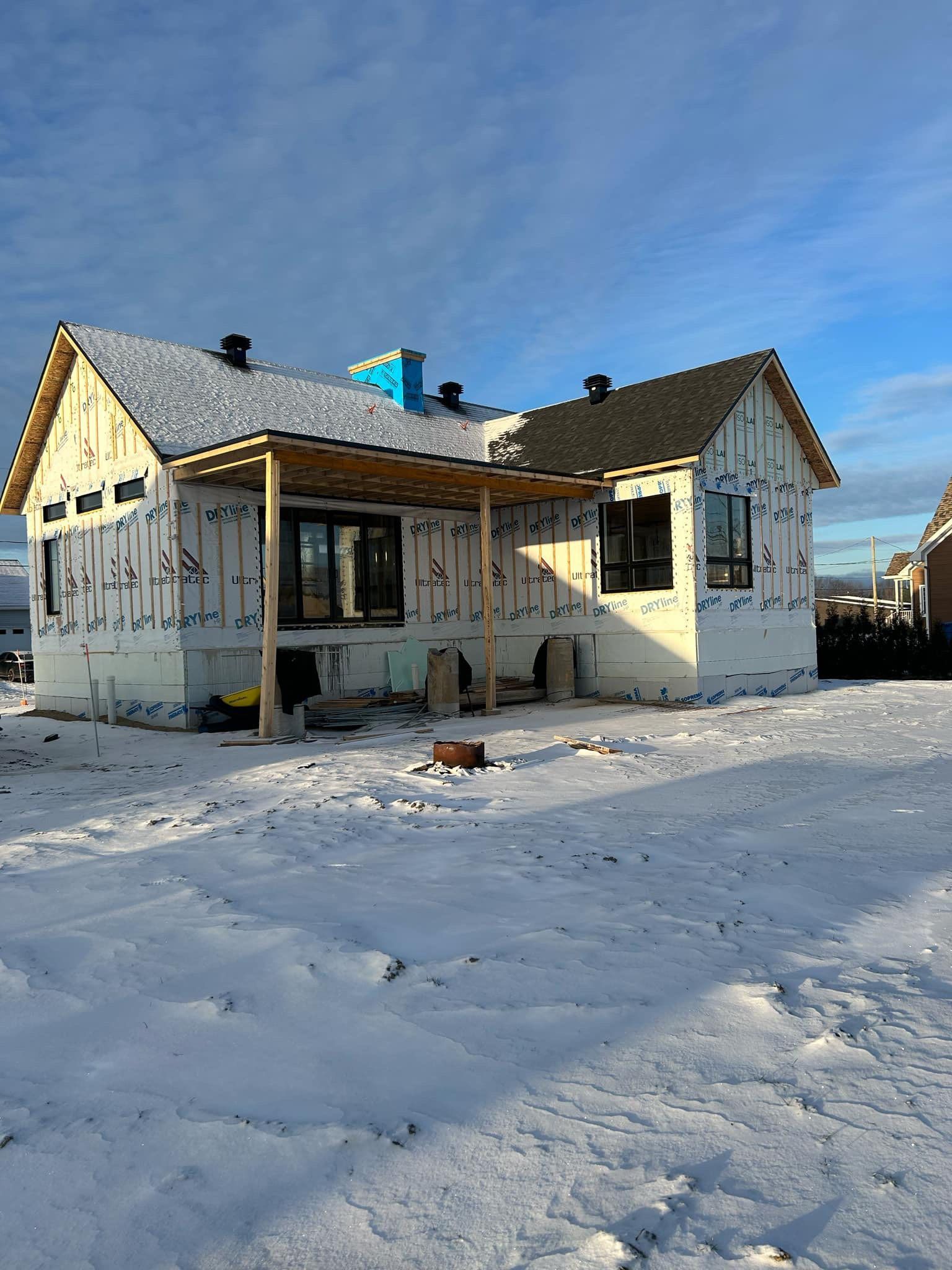Une maison en construction, aux murs blancs isolés et au toit sombre, se dresse dans un paysage enneigé sous un ciel bleu.