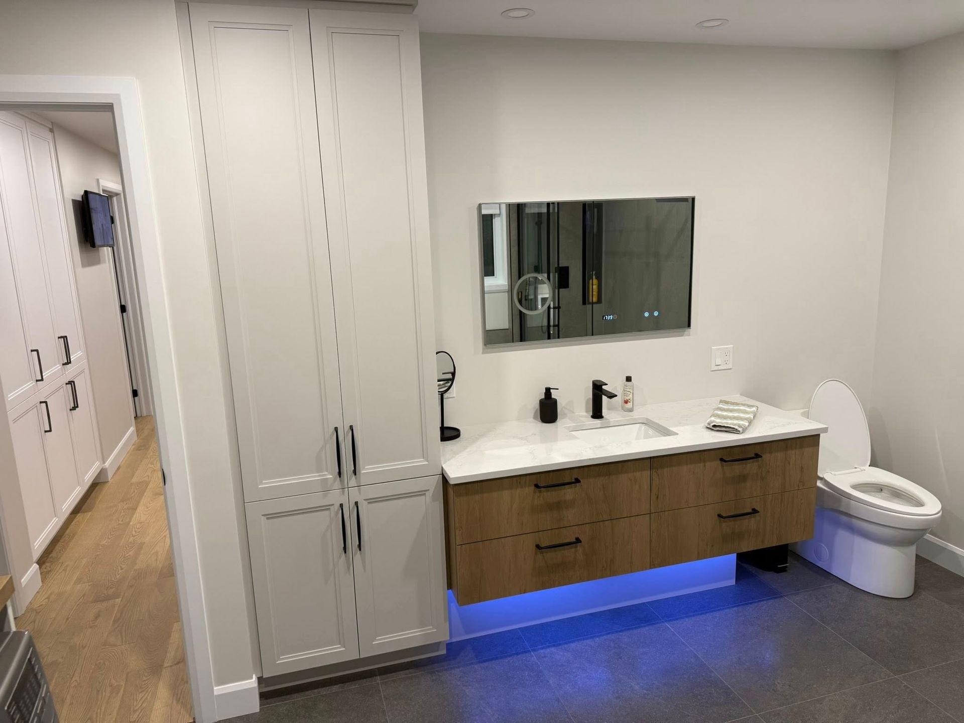 Salle de bains moderne avec meuble-lavabo suspendu en bois clair, armoire de rangement blanche haute, miroir mural et toilettes avec éclairage bleu.