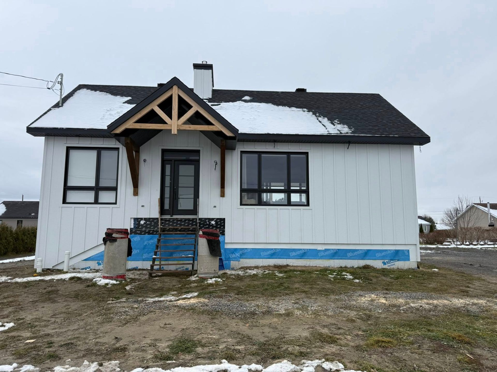Une maison neuve avec un revêtement extérieur blanc, des fenêtres à cadre noir et un porche en bois à pignon, située sur un terrain enneigé.