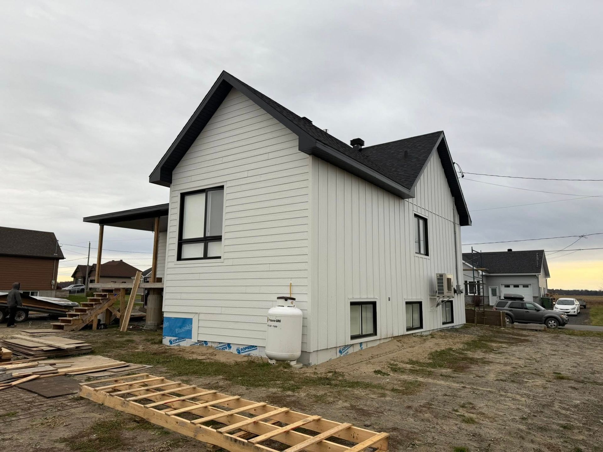 Une maison blanche en construction, avec un toit noir, un bardage noir et une bonbonne de propane, sur un terrain de gravier sous un ciel nuageux.