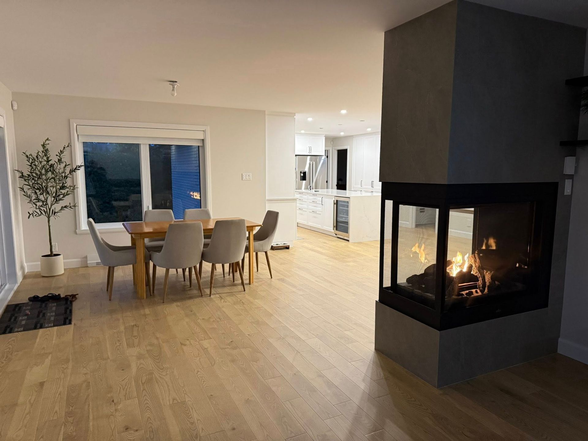 Salle à manger moderne avec une table en bois clair, des chaises grises et une élégante cheminée vitrée à trois faces sur un pilier en béton.