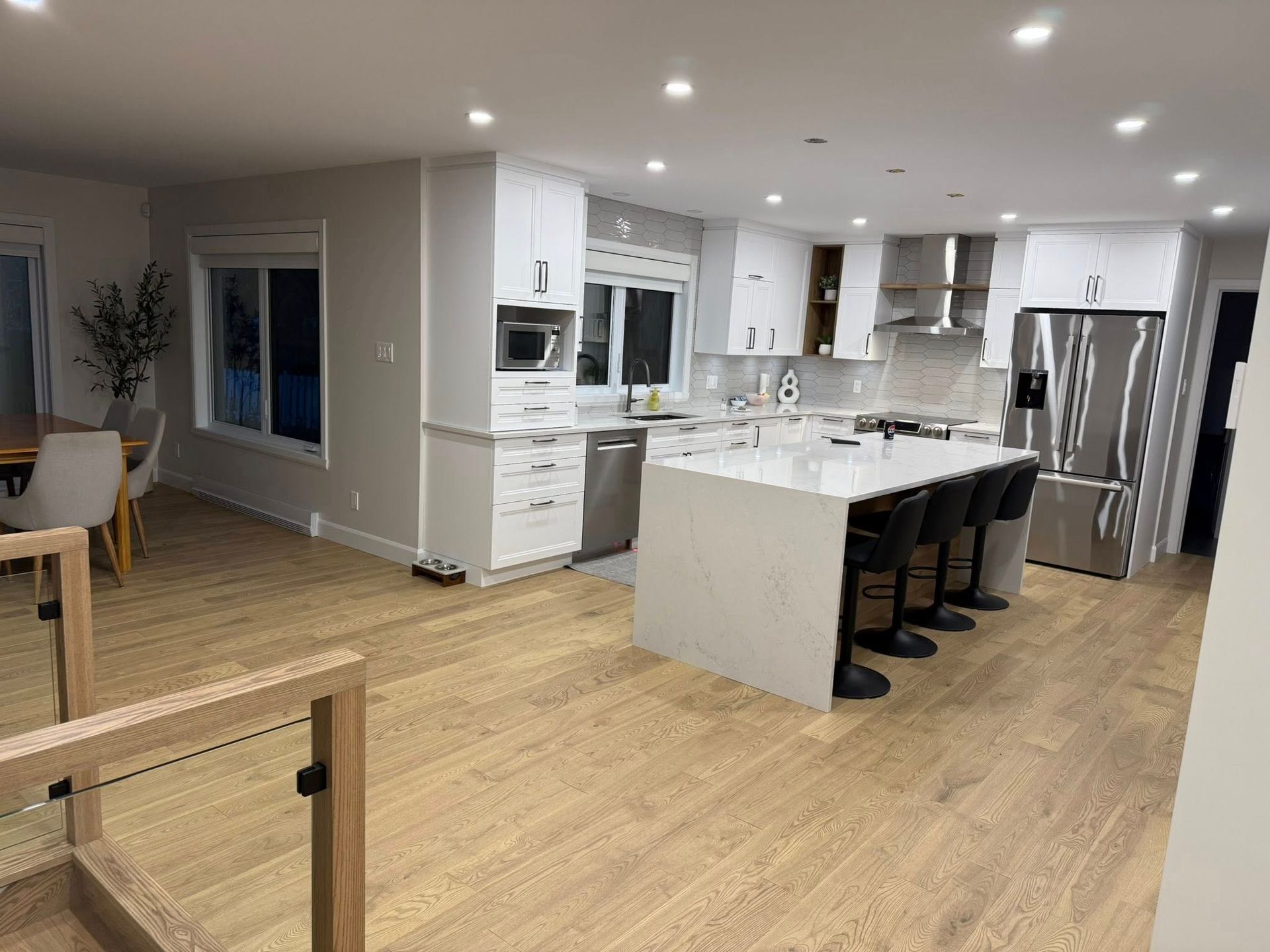 Une cuisine moderne à aire ouverte avec des armoires blanches, un grand îlot avec tabourets de bar, un plancher en bois clair et des luminaires encastrés.