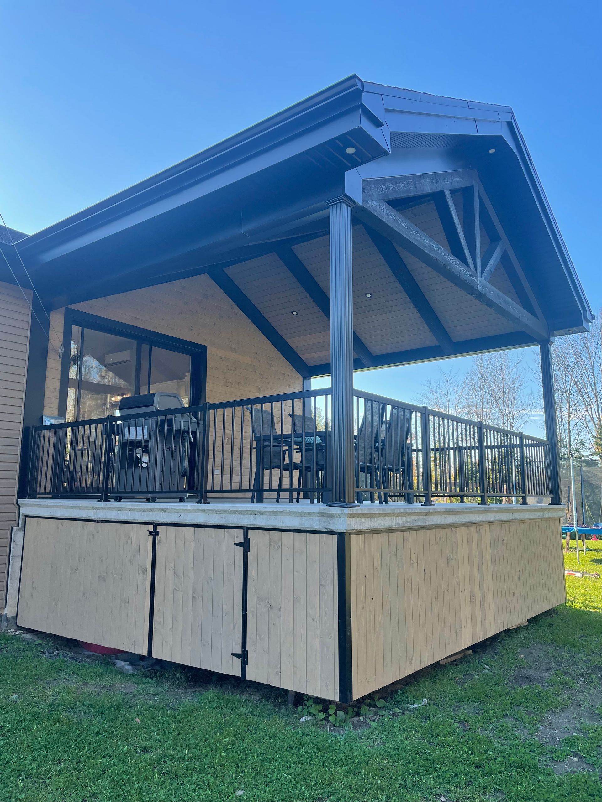 Une terrasse surélevée en bois avec une structure et un toit noirs, comprenant une base en contreplaqué fermée et une rambarde en métal noir.