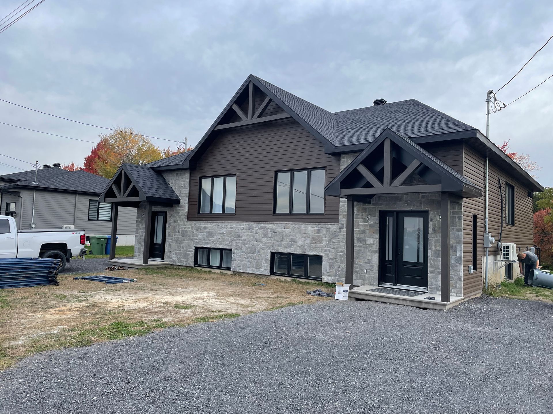 Une maison à deux étages avec un niveau inférieur en placage de pierre, un revêtement extérieur foncé et deux entrées proéminentes à pignon, située sur un terrain en gravier.
