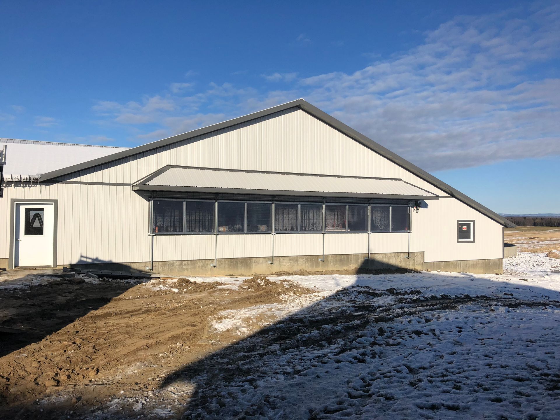 Un bâtiment en métal blanc avec une rangée de fenêtres et un toit gris, se détachant sur un ciel bleu avec un sol enneigé et boueux au premier plan.