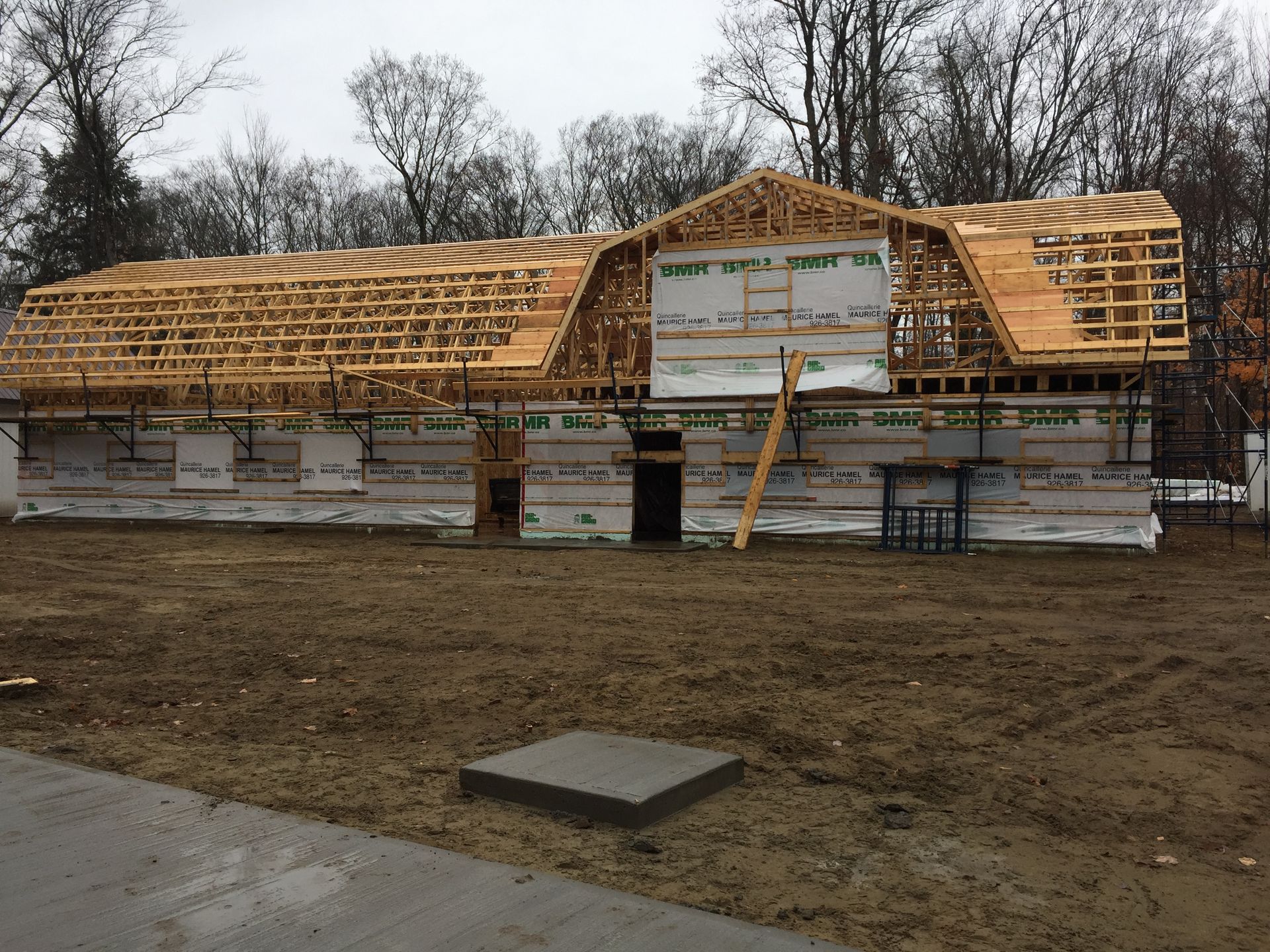 Un bâtiment partiellement construit, avec une charpente de toit en bois et un revêtement extérieur en membrane pare-vapeur, situé sur un terrain en terre battue dans une zone boisée.