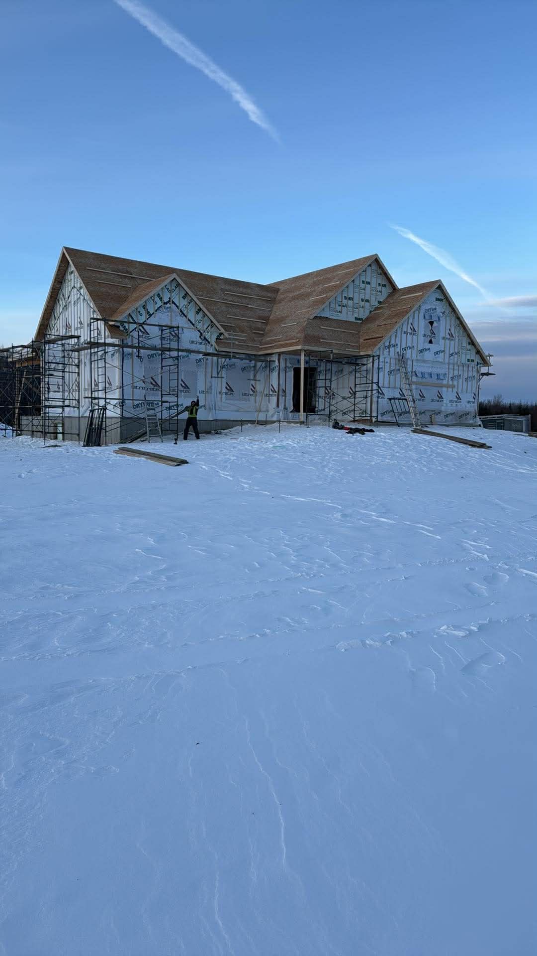 Une maison en construction, avec un revêtement extérieur blanc et un toit en bois, se dresse au milieu d'un champ enneigé sous un ciel bleu.