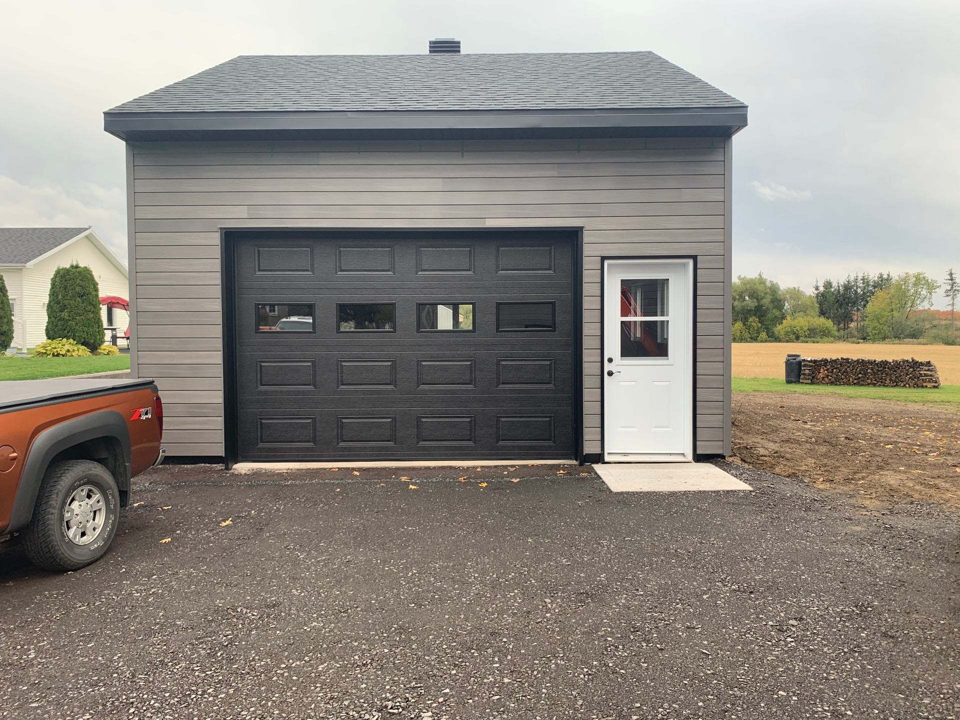 Un garage gris indépendant avec une porte sectionnelle noire et une porte d'entrée latérale blanche, situé sur un terrain gravillonné près d'un champ.