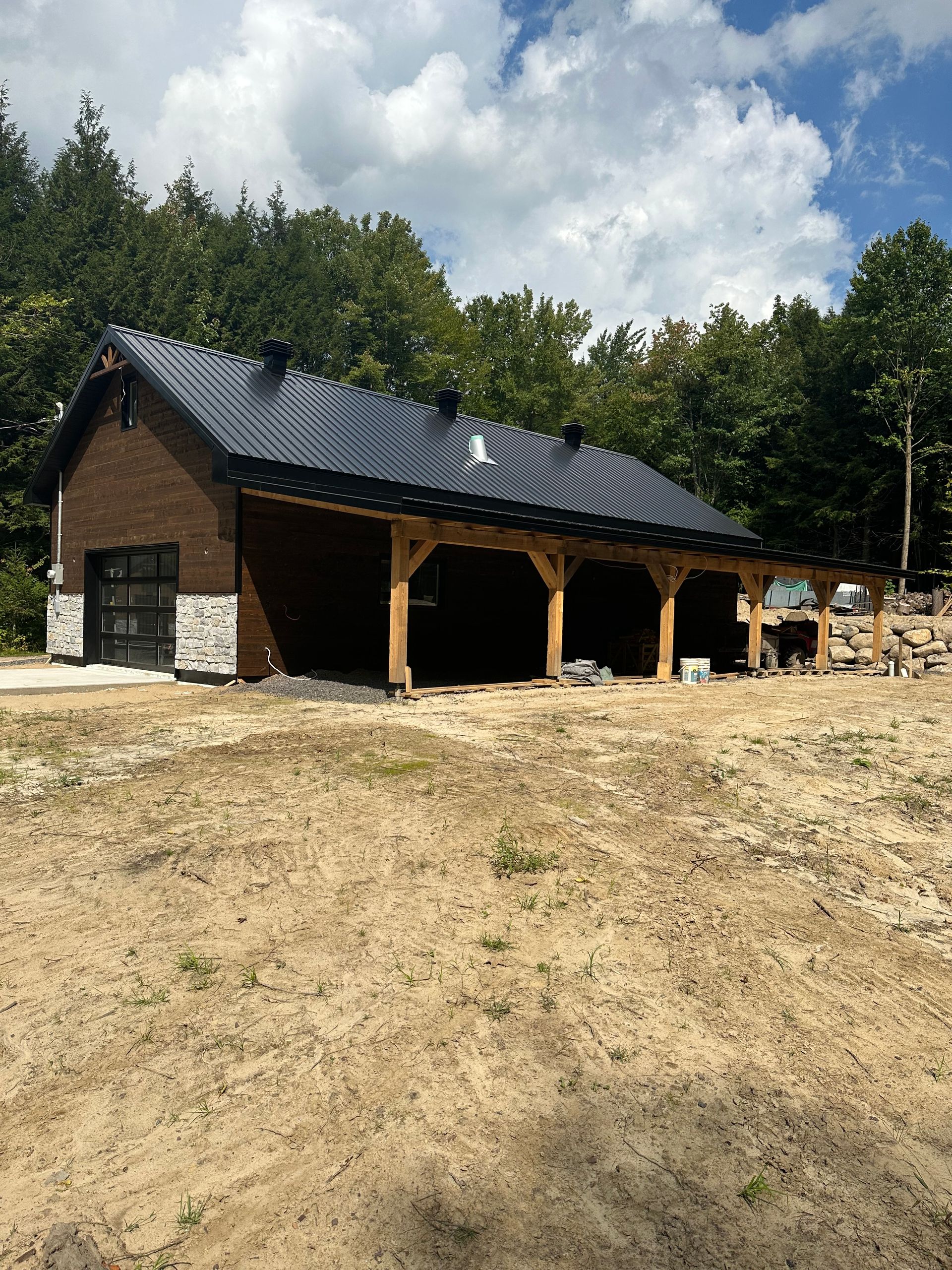 Une grange en bois sombre et en pierre, avec un toit en métal noir et une véranda ouverte, se dresse sur un terrain vague près d'une forêt, sous un ciel bleu.