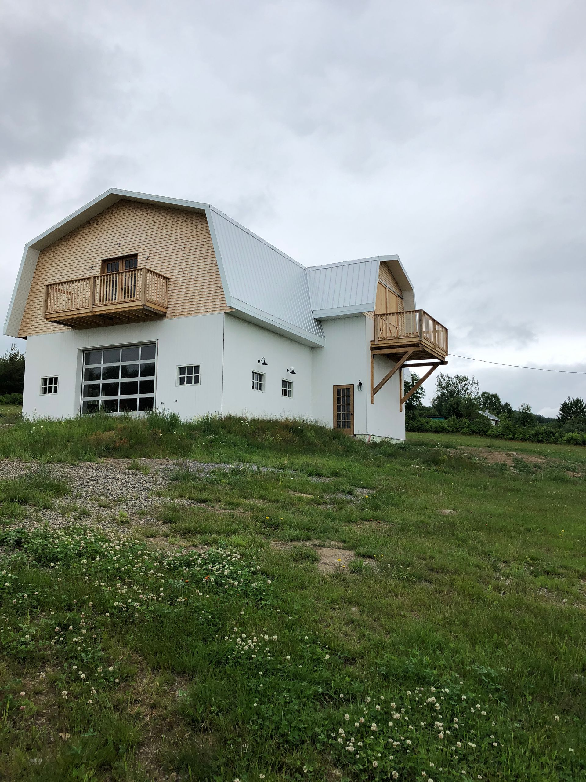 Une grange blanche à deux étages, avec un toit en métal et des balcons en bois, se dresse sur une colline herbeuse sous un ciel nuageux.
