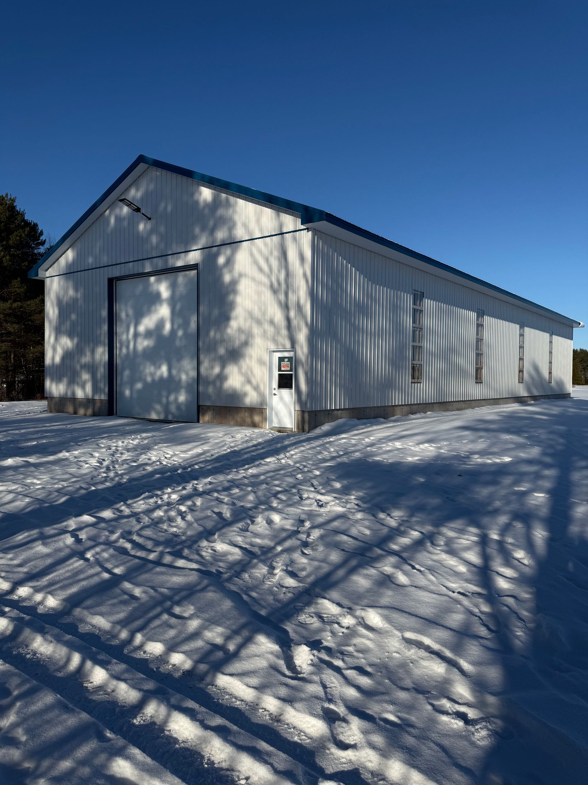 Une grange en métal blanc au toit bleu se dresse dans un champ enneigé sous un ciel bleu dégagé, par une journée d'hiver ensoleillée.